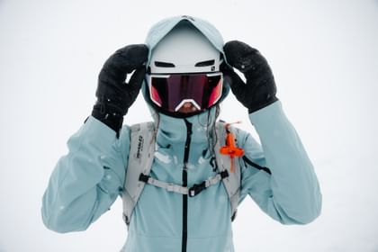 Eine Frau steht mit Skibekleidung, Helm, und Schneebrille frontal und setzt sich die Kapuze auf