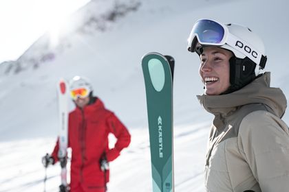 Eine Frau steht mit einem Kästle Ski auf der Piste und lächelt. Im Hintergrund steht ein Mann mit einem Paar Ski in der Hand