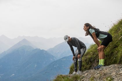 Eine Frau und ein Mann in On Running Bekleidung und Schuhen stehen auf einem Steig in der Bergwelt von Serfaus und atmen durch
