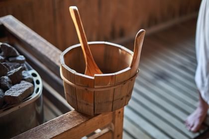 Ein Holzkübel mit Kelle steht neben einem Steinofen in der Sauna zum Aufguss bereit