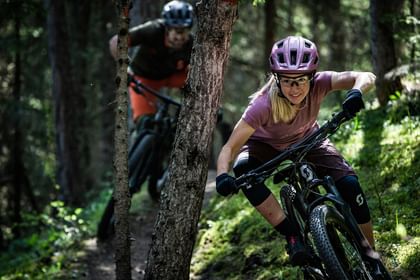 Eine Frau und ein Mann fahren auf einem Fully MTB von Scott einen Trail durch den Wald über Baumwurzeln hinunter