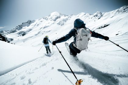 Zwei Personen fahren im Pulverschnee mit Powderski einen Berghang hinunter. Im Hintergrund sind weiße Berggipfel