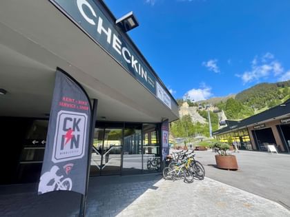 Mountainbikes zum Ausleihen stehen vor dem Geschäft. Im Hintergrund sieht man die Seilbahn Komperdell und das Bergpanorama von Serfaus