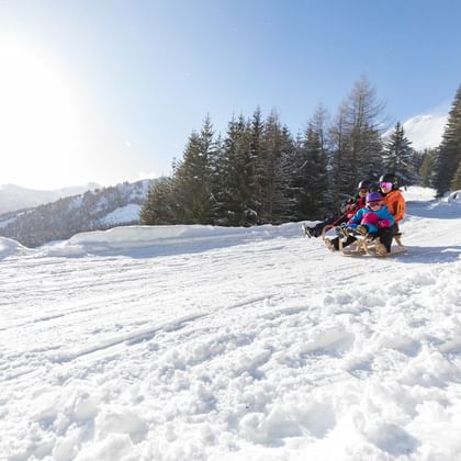 Eine Frau mit Kind und ein weiteres Kind rodeln auf der Rodelbahn in Serfaus-Fiss-Ladis bei strahlend blauen Himmel die Rodelbahn hinunter.