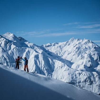 Zwei Skifahrer geben sich am Gipfel eine High-Five im Hintergrund ist ein hochalpines Bergpanorama