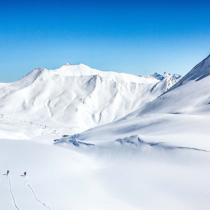 Eine Gruppe von Snowboardern fährt im Masner im Pulverschnee. Im Hintergrund ist ein winterliches Bergpanorama mit tief verschneiten Berghängen und Gipfeln und blauem Himmel im Skigebiet Serfaus-Fiss-Ladis 