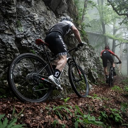 Zwei Männer fahren mit Fully Mountainbikes von Scott einen Trail durch den Wald über Baumwurzeln in dreckiger Bikebekleidung hinunter