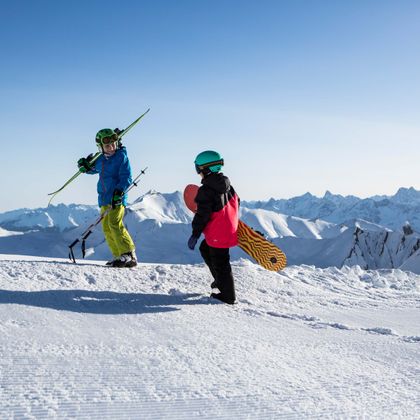 Ein Junge mit Ski und ein Mädchen mit einem Snowboard stehen auf einer Piste in Serfaus und schauen in die weiß verschneite Berglandschaft 