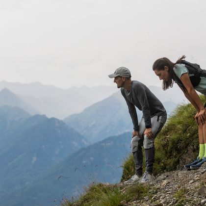 Eine Frau und ein Mann in On Running Bekleidung und Schuhen stehen auf einem Steig in der Bergwelt von Serfaus und blicken in die Landschaft