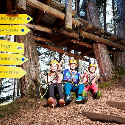 Eine Gruppe von Kindern sitzt vor den Routenschildern im X-Trees Waldseilpark mit ihrer Kletterausrüstung