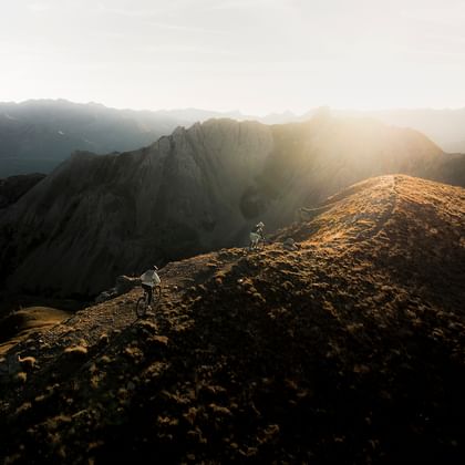 Zwei Fahrradfahrer biken im Sonnenuntergang auf einem Trail am Berggrat. Im Hintergrund sieht man die Berge im goldenen Licht