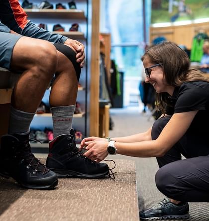 Perfekt sitzende Schuhe mit unserem Happy Feet Bootfitting by Patscheider Eine Verkäuferin hilft einem Kunden beim Anziehen der individuell angepassten Wanderschuhe im Sportgeschäft in Serfaus