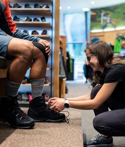 Eine Verkäuferin hilft einem Kunden beim Anziehen der individuell angepassten Wanderschuhe im Sportgeschäft in Serfaus