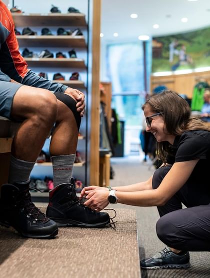 Eine Verkäuferin hilft einem Kunden beim Anziehen der individuell angepassten Wanderschuhe im Sportgeschäft in Serfaus
