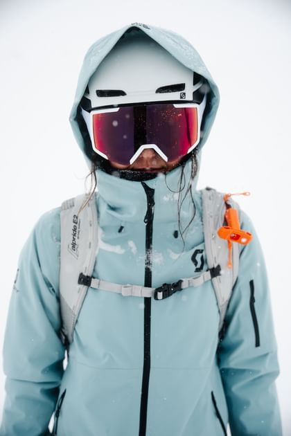 Eine Frau steht mit Skibekleidung, Helm, und Schneebrille frontal. Ihre Haare sind von der Kälte vereist und sie blickt in die Kamera