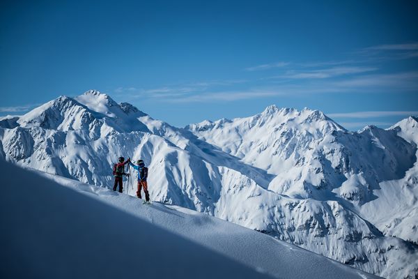 Zwei Skifahrer geben sich am Gipfel eine High-Five im Hintergrund ist ein hochalpines Bergpanorama
