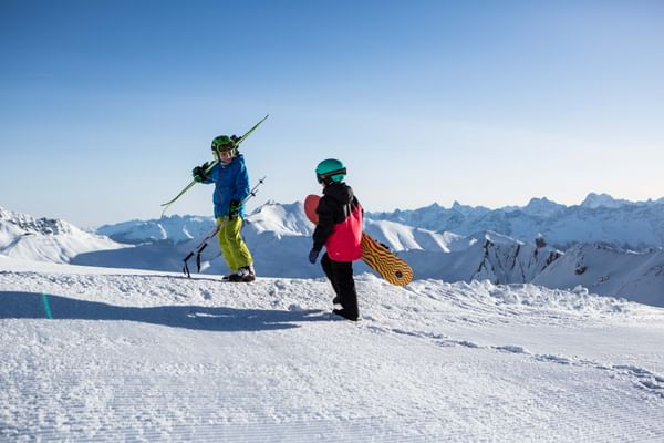 Ein Junge mit Ski und ein Mädchen mit einem Snowboard stehen auf einer Piste in Serfaus und schauen in die weiß verschneite Berglandschaft 