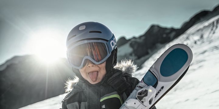 Ein Junge hält im Skigebiet auf der Piste im Sonnenschein Ski in den Armen und streckt die Zunge raus