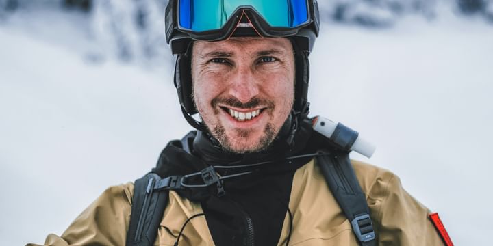 Marcel Hirscher lächelt nach einer Abfahrt im Pulverschnee in den Bergen in einer Mountain Studio Jacke und Helm und Brille zufrieden in die Kamera